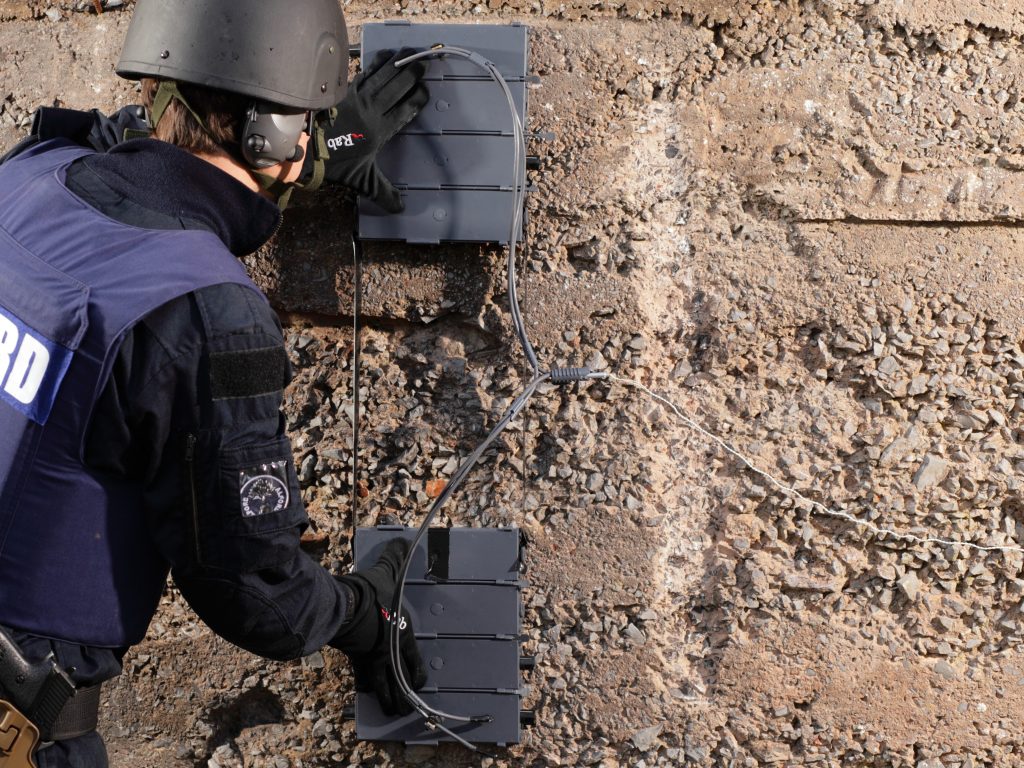 An operator deploying Universal Demolition Block (UDB) charges against a wall